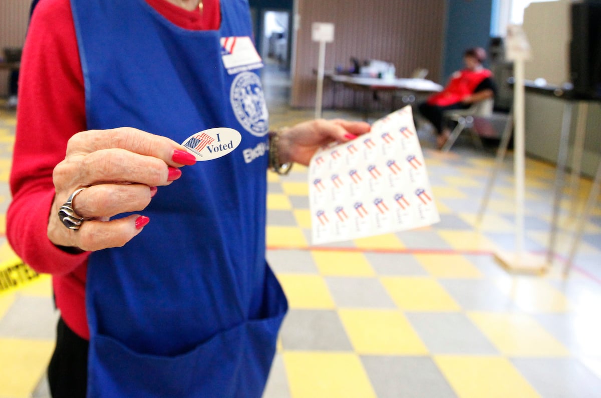 Volunteer seen from neck down, with red nails, handing out I Voted stickers inside a polling place.