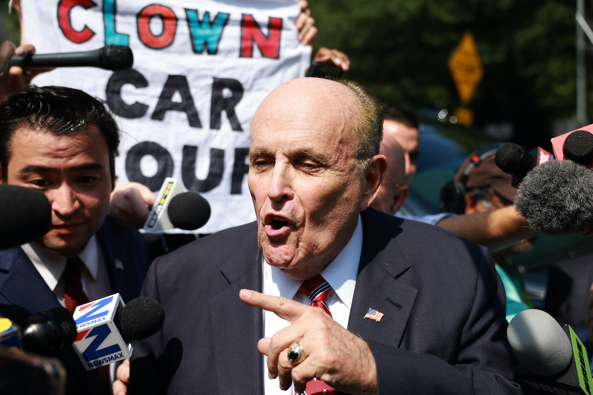 Rudy Giuliani speaks to the media after leaving the Fulton County jail. A sign held behind him reads "clown car coup."