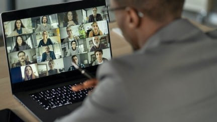 A young Black man seen from behind, on a video call on a laptop with a dozen different people.