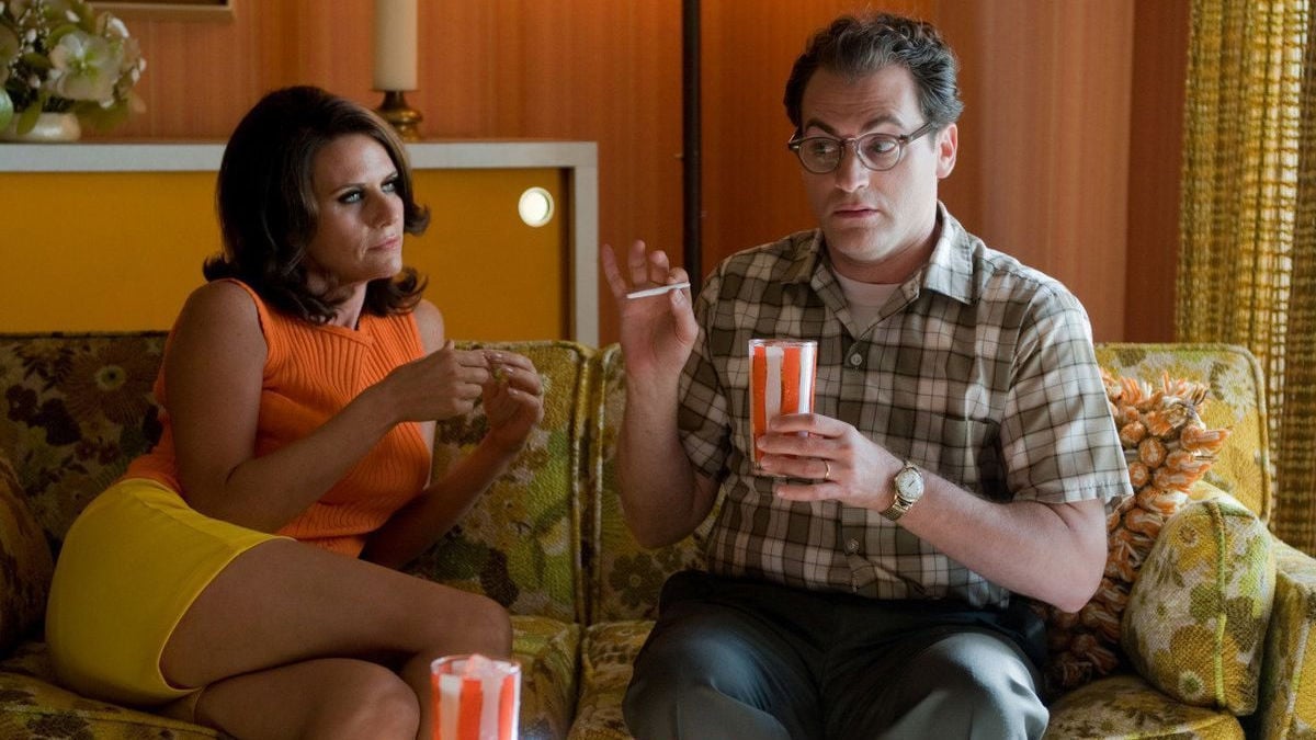 Amy Landecker and Michael Stahlbarg in a scene from 'A Serious Man.' They are both in 1960s period clothing. Landecker is a white Jewish woman with shoulder-length dark hair wearing an orange sleeveless sweater and yellow shorts. Stahlbarg is a white Jewish man with brown hair and a receding hairline wearing black rimmed glasses, a short-sleeved green plaid shirt, and navy blue pants. They are sitting beside each other on a green floral print couch sharing a joint. 