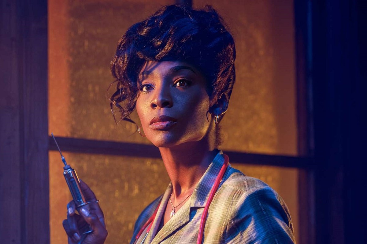 Image of Angelica Ross in a scene from 'American Horror Story.' She is a Black trans woman with her long dark hair pulled up with wavy tendrils hanging down one side of her face. She's wearing a light-colored plaid collared shirt with a stethoscope around her shoulders and is holding a syringe. She's looking at something in the distance. 