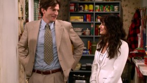 Ashton Kutcher and Mila Kunis in a scene from Netflix's 'That 90s Show.' Kutcher is a white man with shaggy hair wearing a suit and tie. Kunis is a white woman with long dark hair wearing a white suit. They are standing in a kitchen as they look at each other, smiling.