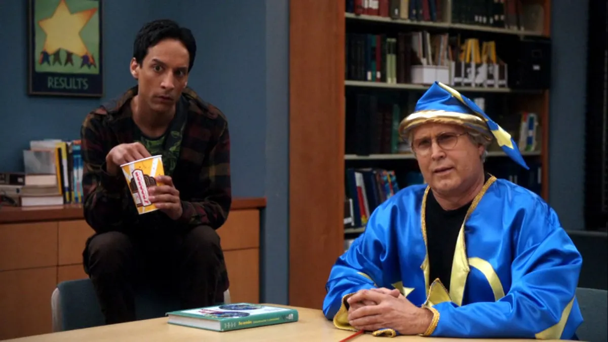 Danny Pudi (Abed) and Chevy Chase (Pierce) sit at a table in a still from 'Community'.