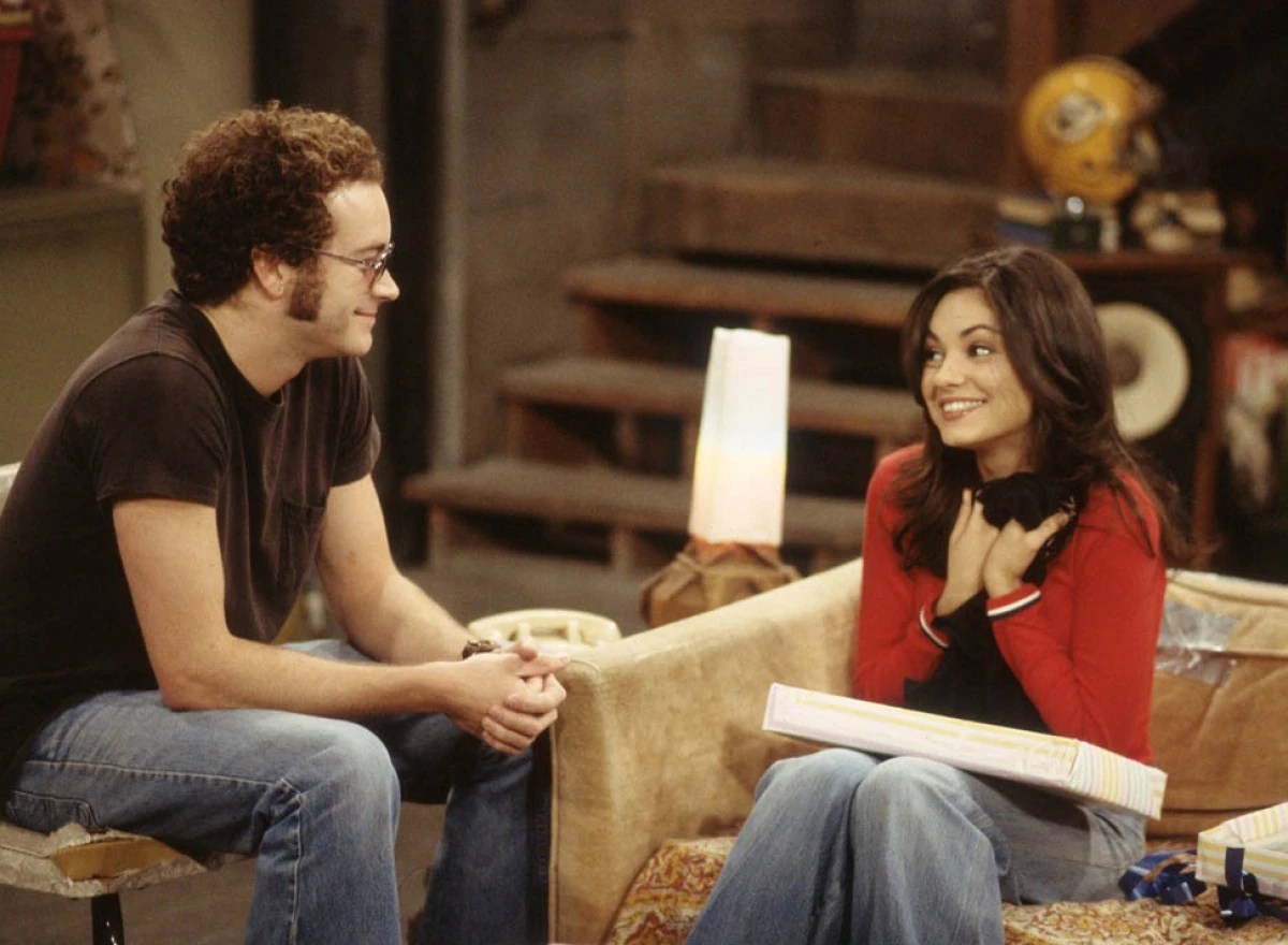 Danny Masterson and Mila Kunis in a scene from FOX's 'That 70s Show." Masterson is a white teenage boy with curly reddish hair sitting in a chair in a furnished basement wearing a brown t-shirt and jeans. Kunis is a white teenage girl with long brown hair sitting on a couch wearing a red cardigan, black shirt, and jeans. They are looking at each other and smiling.