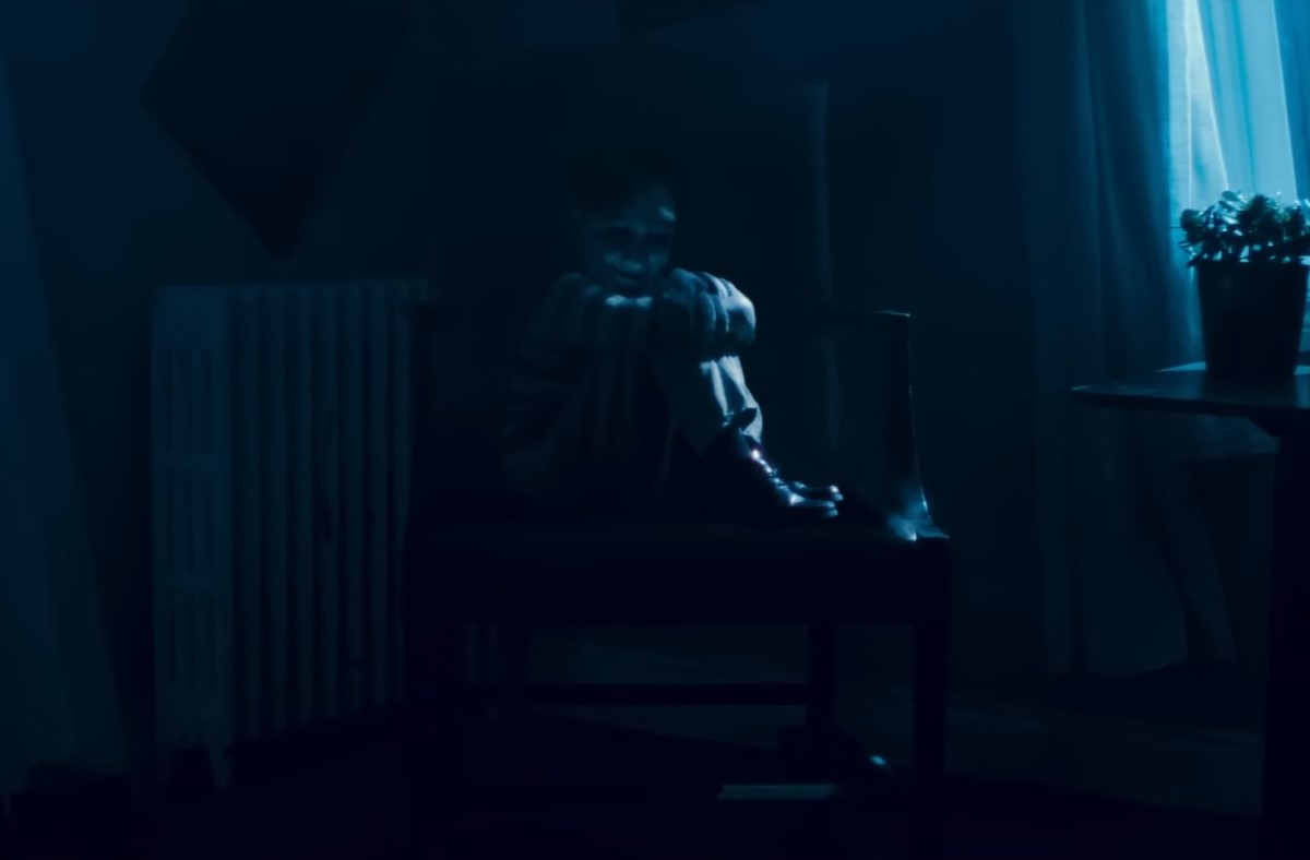 A scene from 'Dear David.' A creepy little boy sits turned to the side on a large chair with his knees pulled up to his chest. He's looking toward the camera smiling creepily in the shadows. 