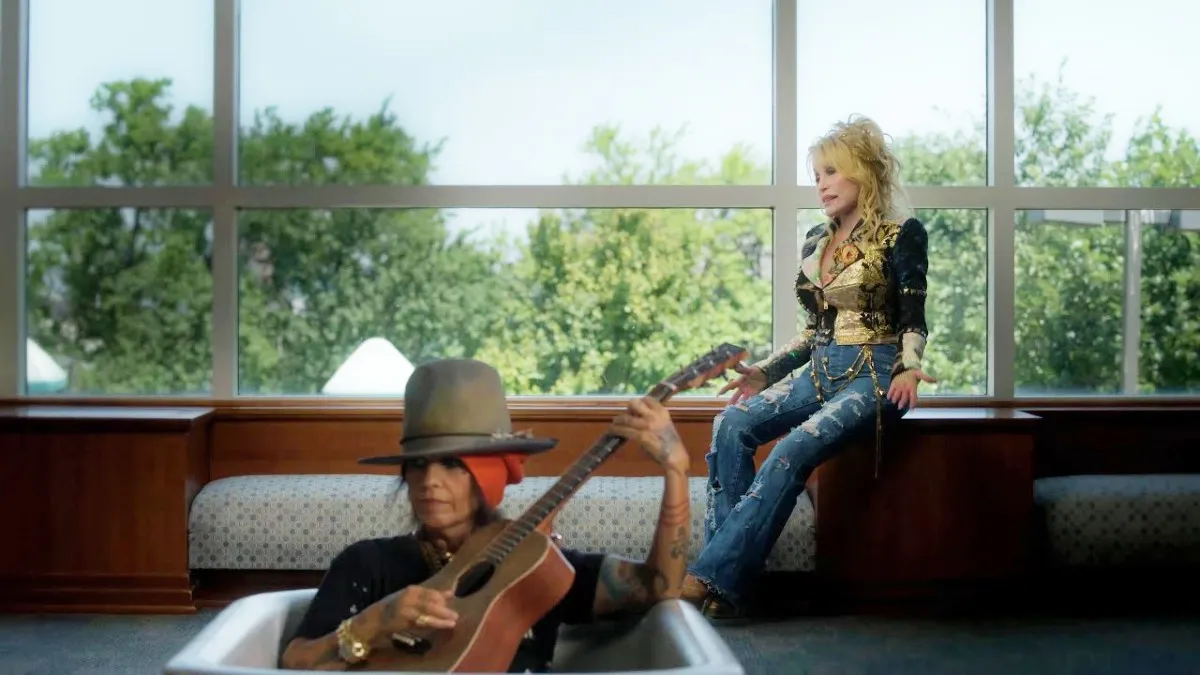 A woman sings while looking out of a window while another woman plays guitar in a bathtub in music video for Dolly Parton's "What's Up?"