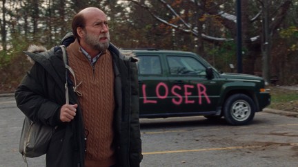 Nicolas Cage stands in front of a car spraypainted with the word 