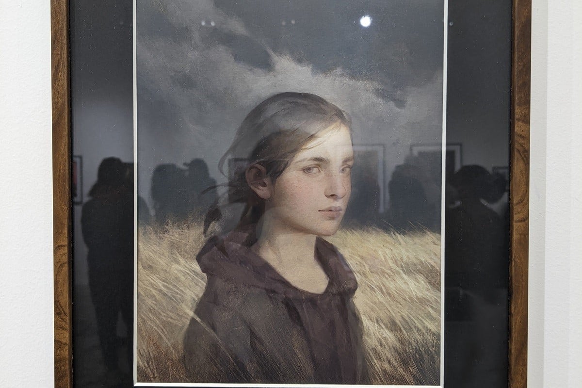 Image of the framed painting "Ellie's World" by Betty Jiang. It's a painting of Ellie from 'The Last of Us' (white teenage girl with brown hair in a ponytail wearing a dark hoodie under a green jacket) from the shoulders up glancing sideways at the viewer with a serious expression. She's standing in a field of tall yellow grass blowing in the wind in front of a dark sky. 