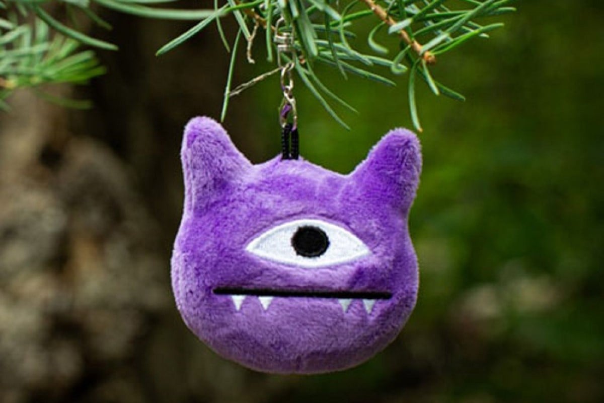Close-up of "Ellie's Monster Keychain." It's a plush, purple monster with cat-like ears, one eye, and 4 sharp teeth. 
