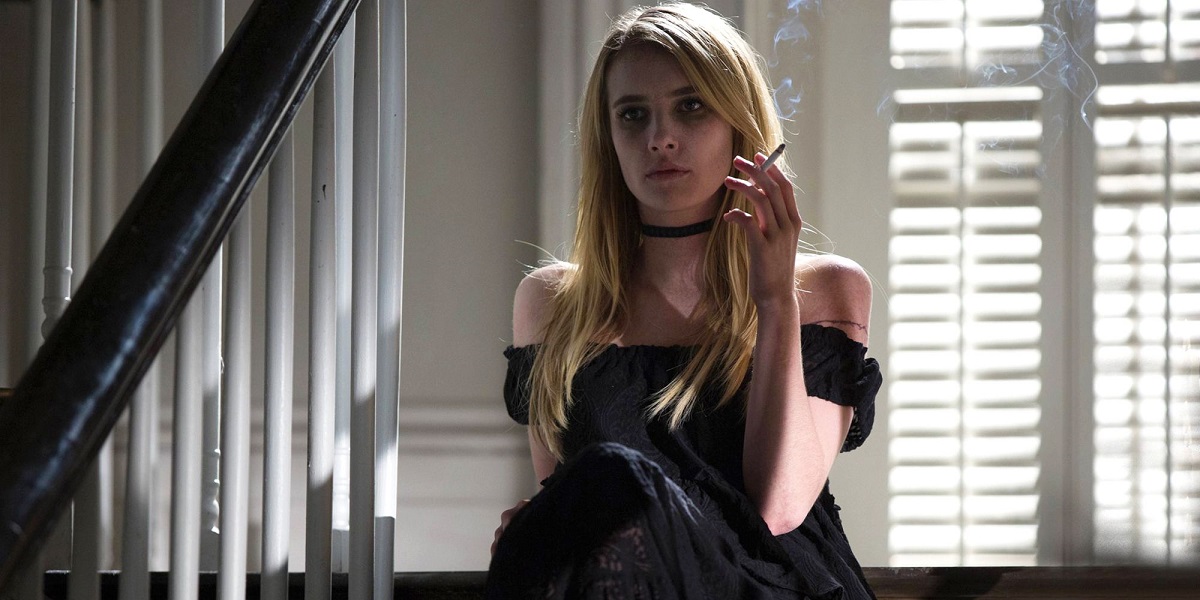 Image of Emma Roberts in a scene from FX's 'American Horror Story.' She is a blonde white cis woman with long straight hair sitting at the top of a staircase in a house. She's wearing a black off-the-shoulder dress and a black choker. Her legs are crossed and she's holding a lit cigarette as she looks straight ahead.