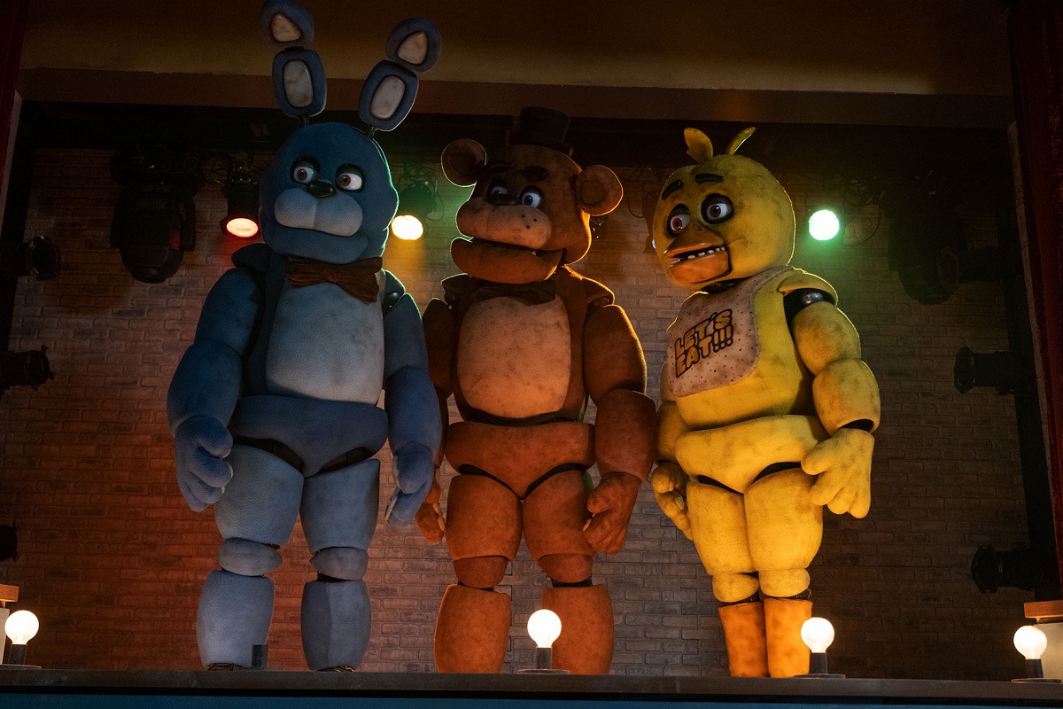 Image of a large animatronic rabbit, bear, and chick in a scene from the film  'Five Nights at Freddy's.' They are standing on a stage and all looking in the same direction. 