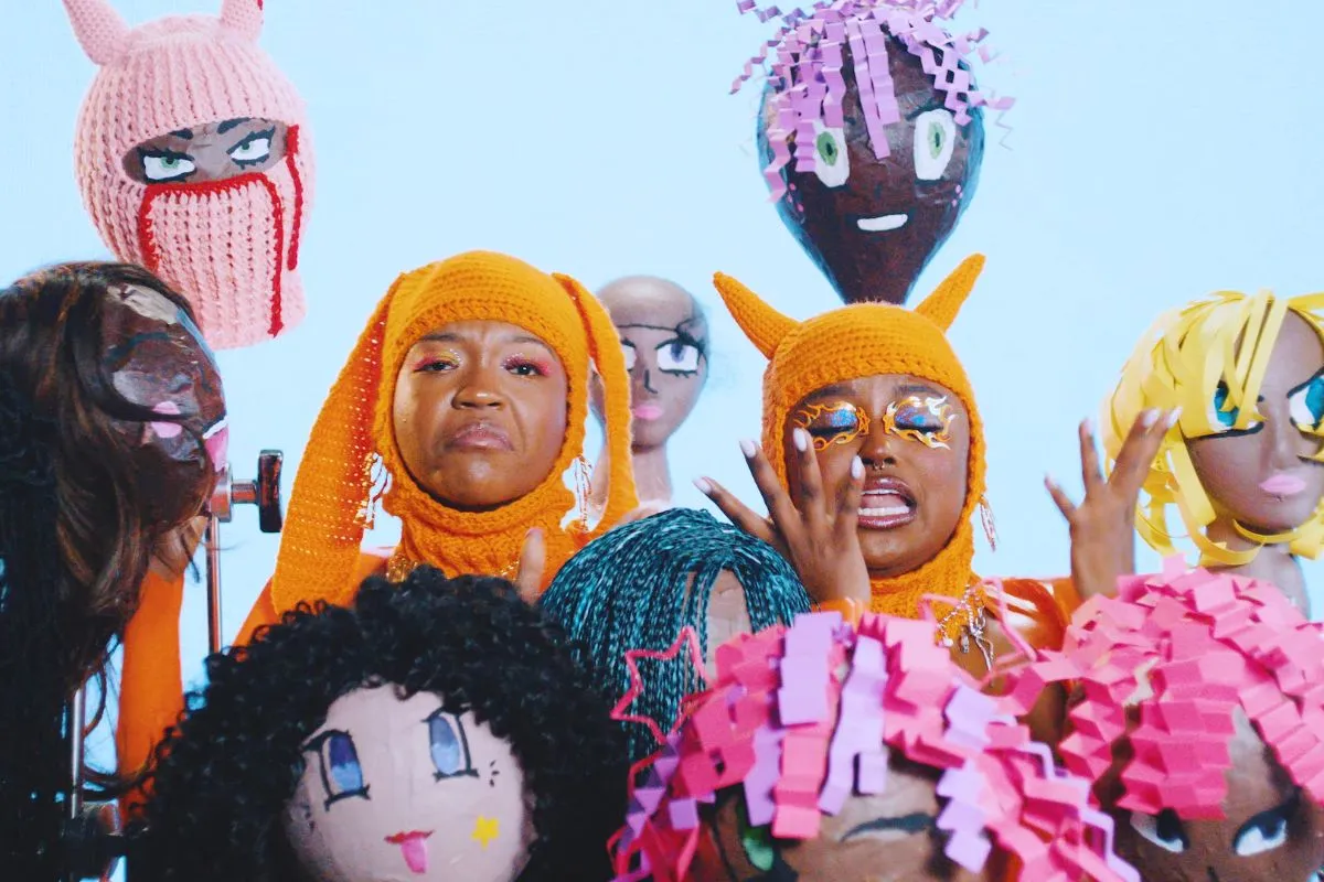 Bobbi Lanea Tyler and Folayan Omi Kunerede of Flyana Boss next to paper machete heads of other Black women in the You Wish music video.