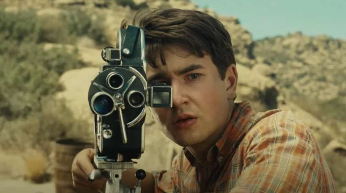 Gabriel LaBelle as Sammy in 'The Fablemans.' He is a white Jewish teenage boy with short shaggy brown hair wearing an orange plaid buttondown. He's standing outside looking through the viewer on an 8 millimeter film camera.