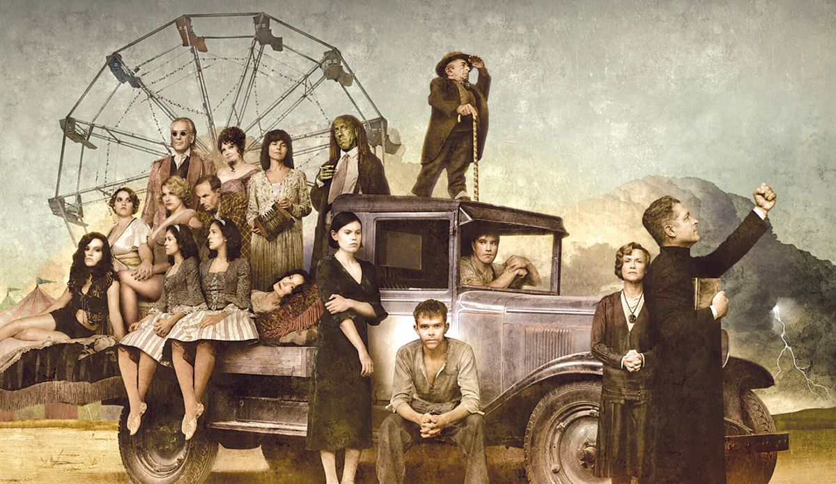 A group of carnies stand around a 1930s pickup with a large ferris wheel in the background in 'Carnivale.'