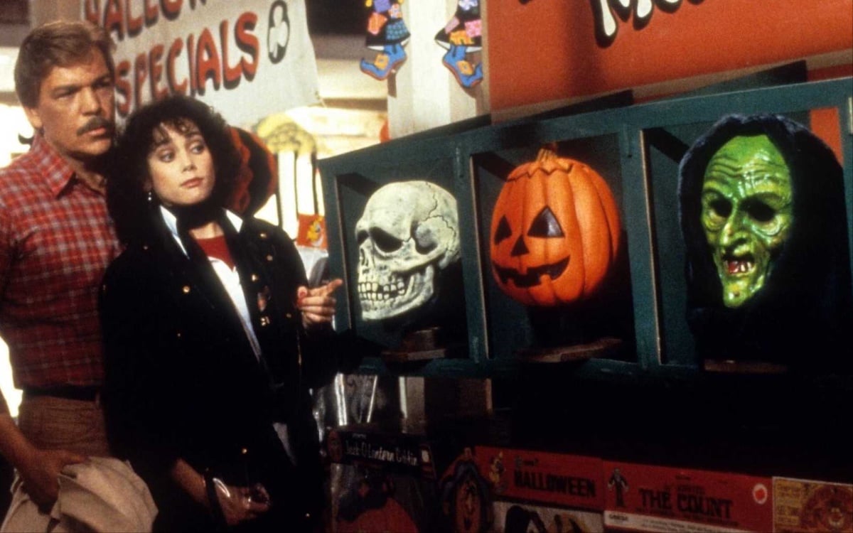 Image of Tom Atkins as Dr. Dan Challis and Stacey Nelkin as Ellie Grimbridge in Halloween III: Season of the Witch. Depicts characters looking at iconic skeleton and pumpkin masks from the film.