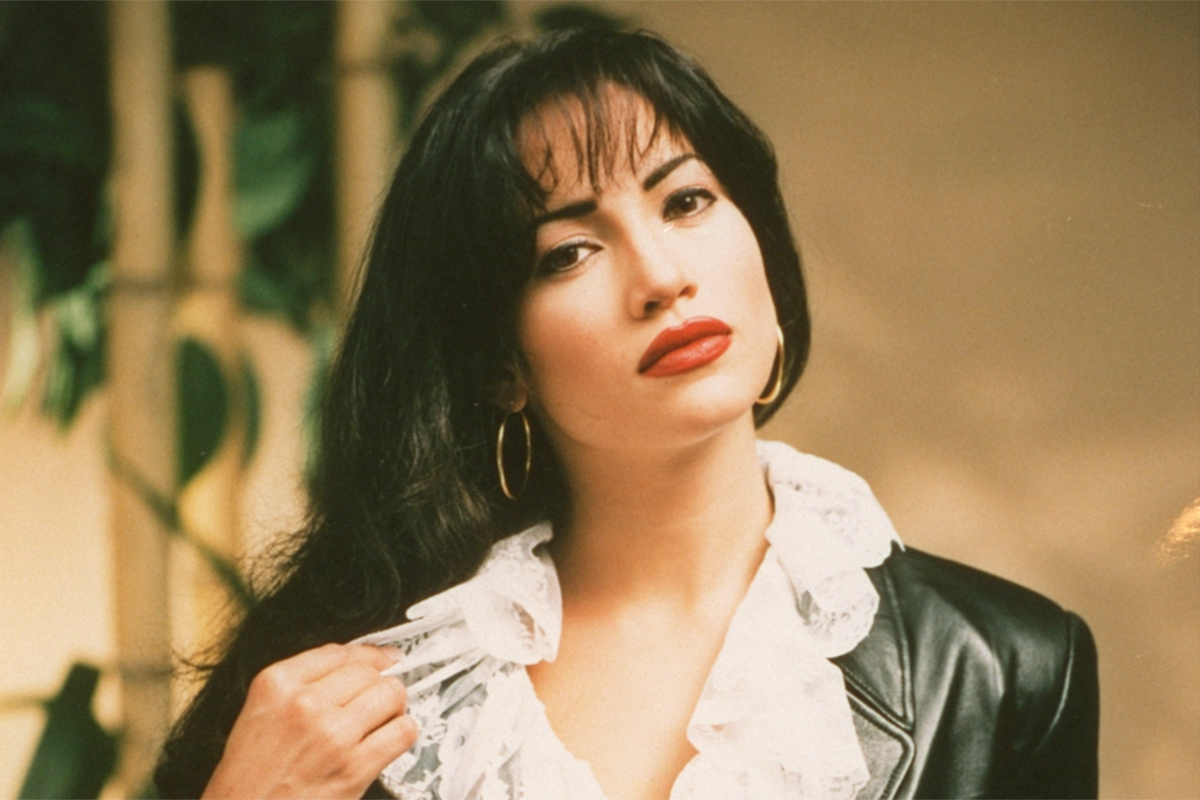 Jennifer Lopez as Selena Quintanilla in the 1997 film 'Selena.' She is a Latina with long black hair and bangs wearing bright red lipstick, silver hoop earrings, and a lacy shirt under a black leather jacket. We see her from the shoulders up as she looks into the camera. 