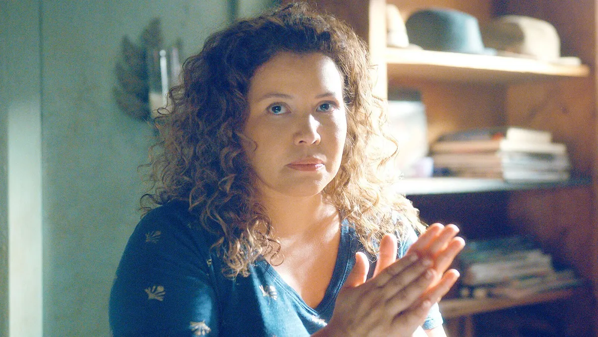 Justina Machado in a scene from Amazon's 'The Horrors of Dolores Roach.' She is a Latina with shoulder-length, curly brown hair wearing a navy blue v-neck long-sleeved sweater with white dandelions on it. She's inside looking seriously at something as she rubs her hands together.