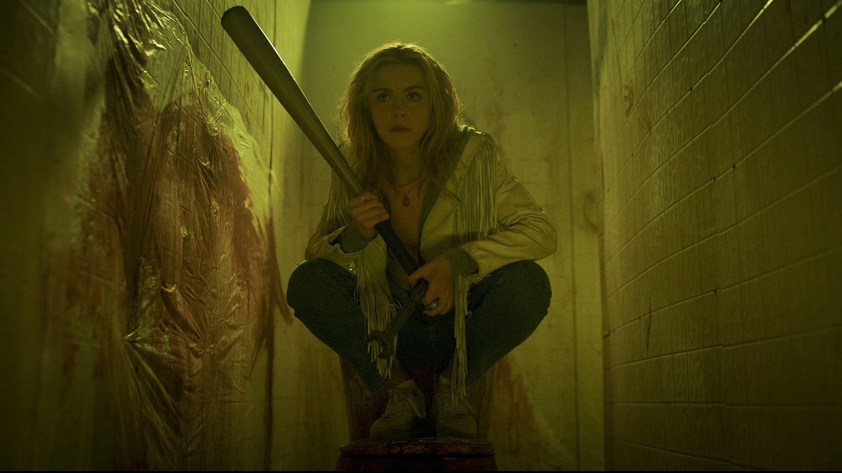 Kiernan Shipka in a scene from Amazon Prime's 'Totally Killer.' She's a white, blonde young woman with long straight hair. She's crouched and wearing a white jacket with fringe over a yellow t-shirt and jeans as she holds a baseball bat in a blood-splattered hallway. 