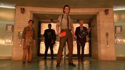 Tom Hiddleston stands in front of three other castmates in a scene from 'Loki' on Disney+. The four (three men and one woman) stand heroically in a large room.