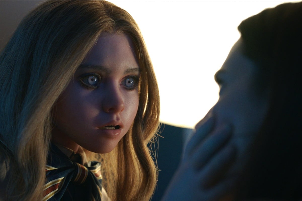 Image of the doll M3GAN (pronounced Megan) in the film of the same name. She is a life-sized doll that looks like a redhead tween girl with light eyes. She's leaning in menacingly while holding Allison Williams' face.