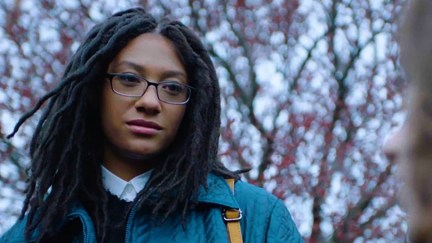 Madeline Grey DeFreece as Carrie in the film 'Tahara.' She is a Black, Jewish teenager with long black dreadlocks wearing black-rimmed glasses and a blue jacket over a black sweater and white buttondown. She's standing outside with budding trees in the background.
