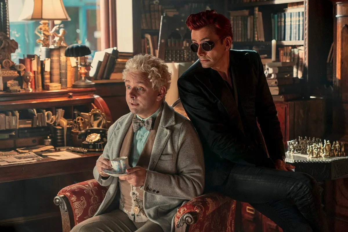 Michael Sheen and David Tennant in a scene from Amazon Prime's 'Good Omens." They are in a bookshop. Sheen is sitting in an armchair holding a cup of tea while Tennant sits on the arm of the chair. Both are white men. Sheen has white, shaggy hair and is wearing a light-colored three-piece suit with a plaid bow tie. Tennant is wearing all black, including sunglasses, and has short red hair in a pompadour.