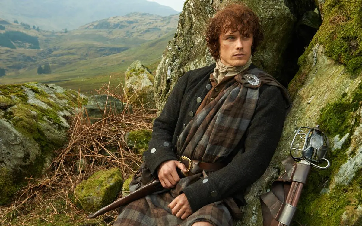 Red haired man wearing a kilt leans against a rock with sprawling landscape behind him in 'Outlander.'