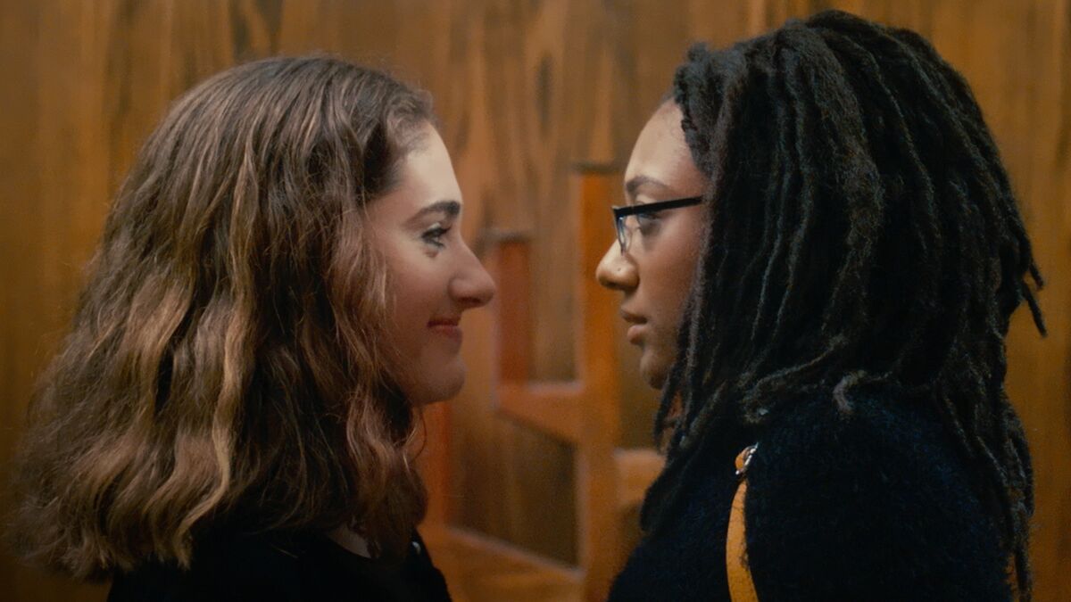 Rachel Sennott as Hannah and Madeline Grey DeFreece as Carrie in the film 'Tahara.' They stand face to face in a synagogue and we see them in profile from the shoulders up. Hannah is a white Jewish teenage girl with brown shoulder-length hair. Carrie is a Black Jewish teenage girl with shoulder-length dreadlocks wearing black-rimmed glasses. 