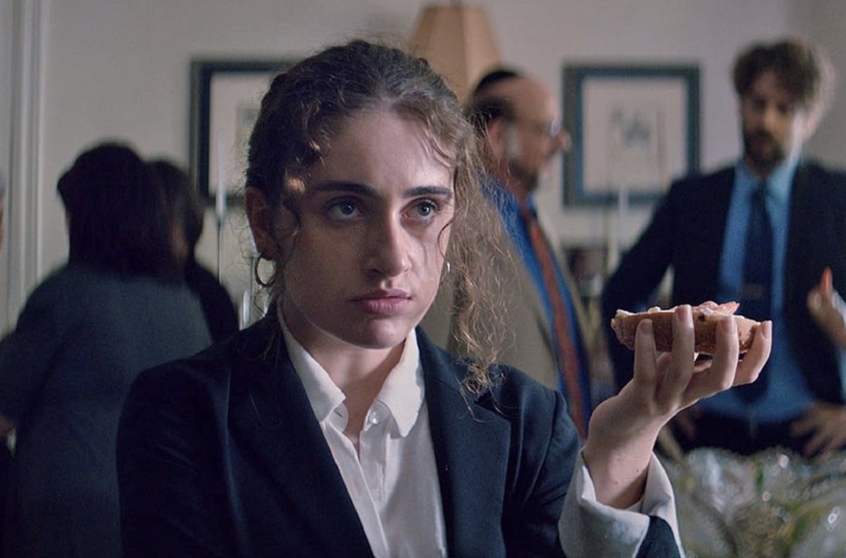 Rachel Sennott as Dani in the film 'Shiva Baby.' She is a white Jewish young woman with her brown curly hair pulled back into a pony tail. She's wearing a black blazer over a white buttondown shirt and holding a bagel in her hand as she stands in the middle of a family gathering.