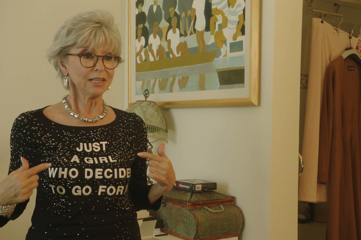 Rita Moreno stands pointing at the black sparkly shirt with the words "Just a Girl Who Decided to Go For It" in a scene from the documentary of the same name. Moreno is an elderly Latina with short, grey hair. 