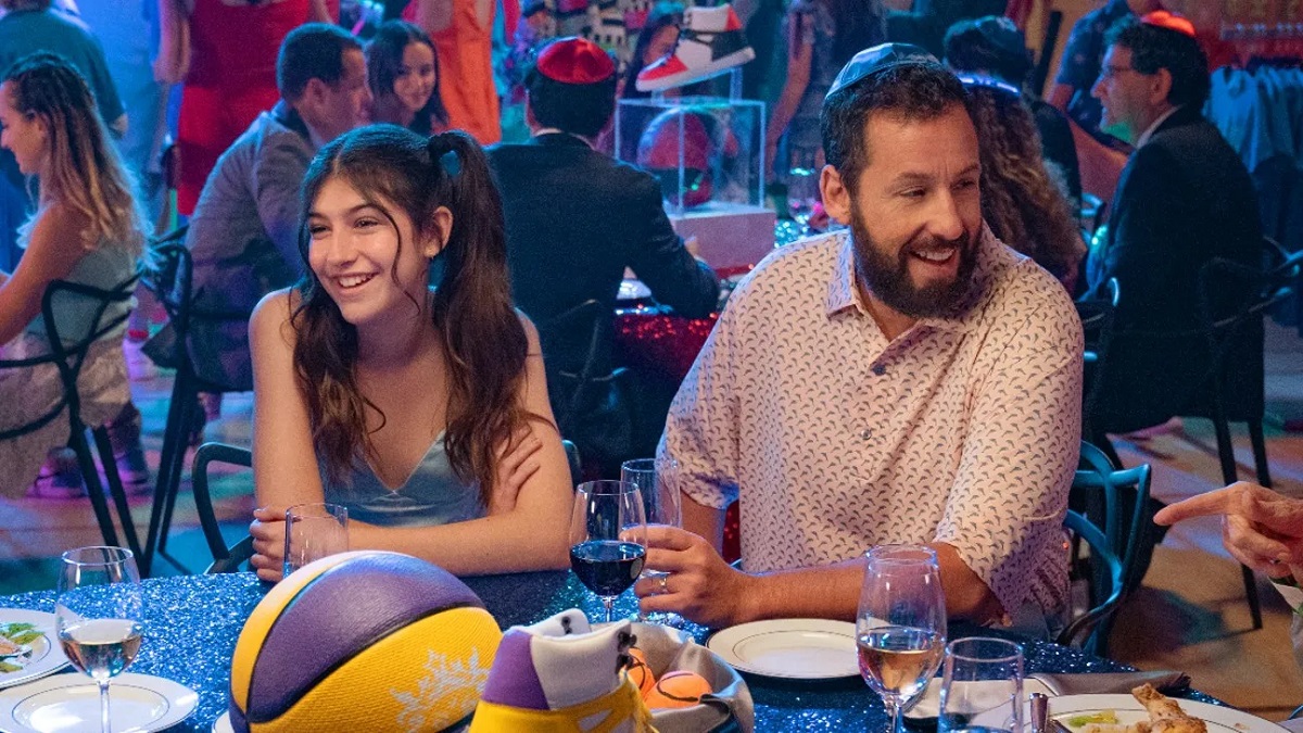 Sunny Sandler as Stacy and Adam Sandler as Danny in a scene from Netflix's 'You Are So Not Invited to My Bat Mitzvah.' They are sitting at a table set for dinner at a party. Stacy is a white Jewish girl with her long, dark hair in pigtails wearing a blue satin tank top. She sits smiling with her arms folded on the table. Danny is a white Jewish man wearing a yarmulke with short dark hair and a beard. He's sitting with one arm on the table holding a glass of wine and smiling wearing a white patterned buttondown.