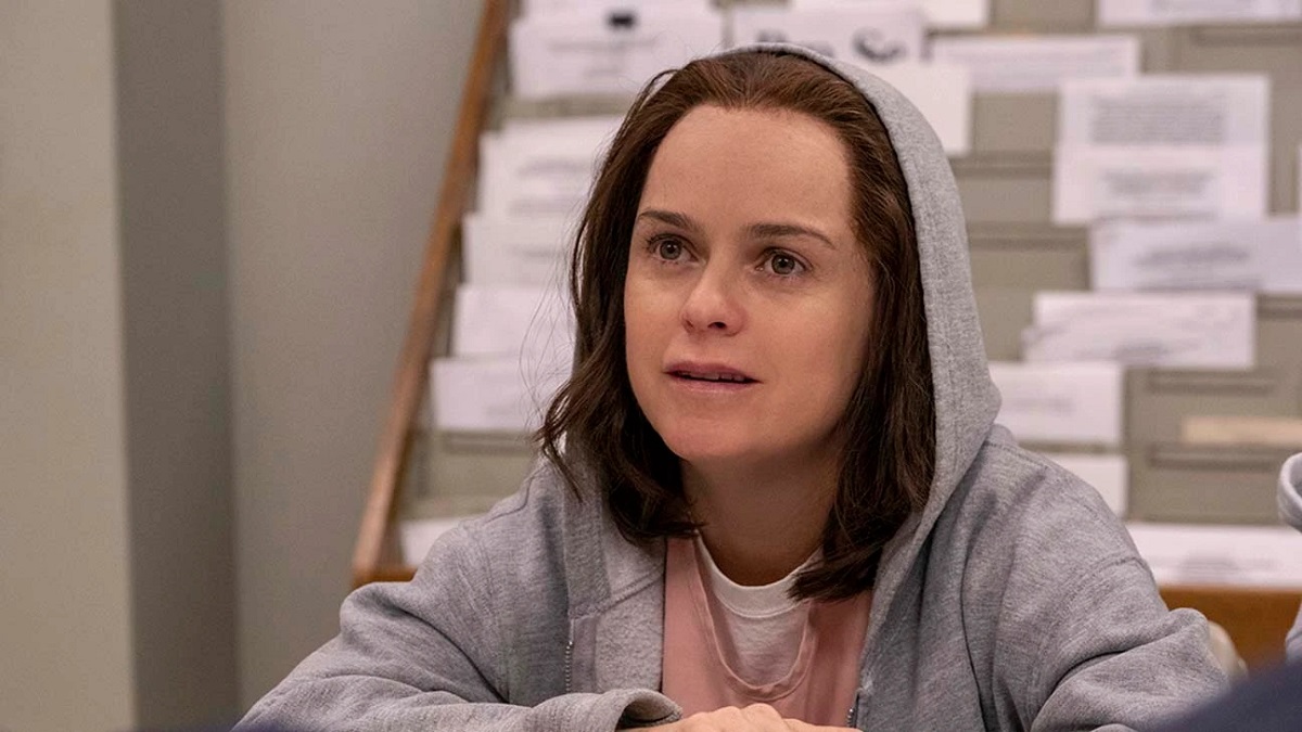 Taryn Manning in a scene from Netflix's 'Orange is the New Black.' She is a white woman sitting in an office with her arms folded on a table. She has shoulder-length brown hair and is wearing a hoodie over her prison scrubs.