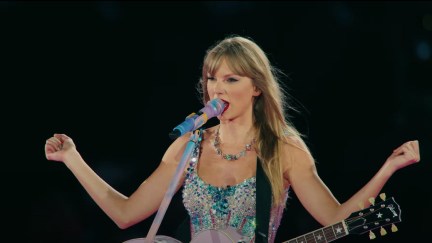 Screencap of Taylor Swift from the trailer for her concert film, 'Taylor Swift: The Eras Tour.' She is a white woman with long blonde hair and bangs wearing a white, sleeveless dress with sparkling stones in different shades of blue on the bodice and straps. She has a guitar hanging from her shoulders and her arms are outstretched as she sings into a microphone.
