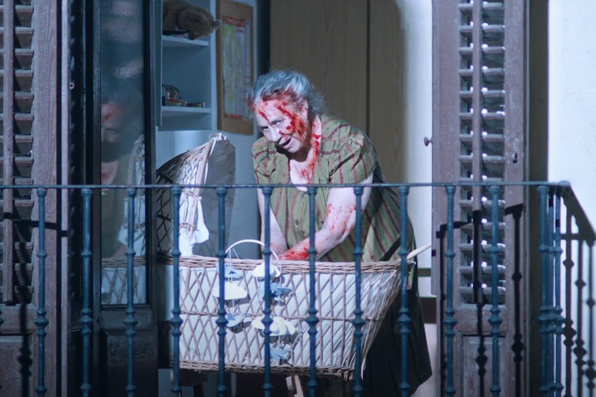 An elderly woman in a scene from 'The Elderly.' She is a white Spanish woman standing on a balcony with her white hair swept up on her head. She's wearing a green short sleeved shirt covered in blood and reaching into a bassinet. 