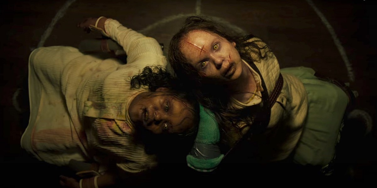Image of Lidya Jewett as Angela and Olivia Marcum as Katherine in a scene from 'The Exorcist: Believer.' Angela is a young Black girl and Katherine is a young white girl. Both are in nightgowns and tied to chairs back-to-back as they look up at the camera. They are possessed and covered in cuts and bloodstains. 