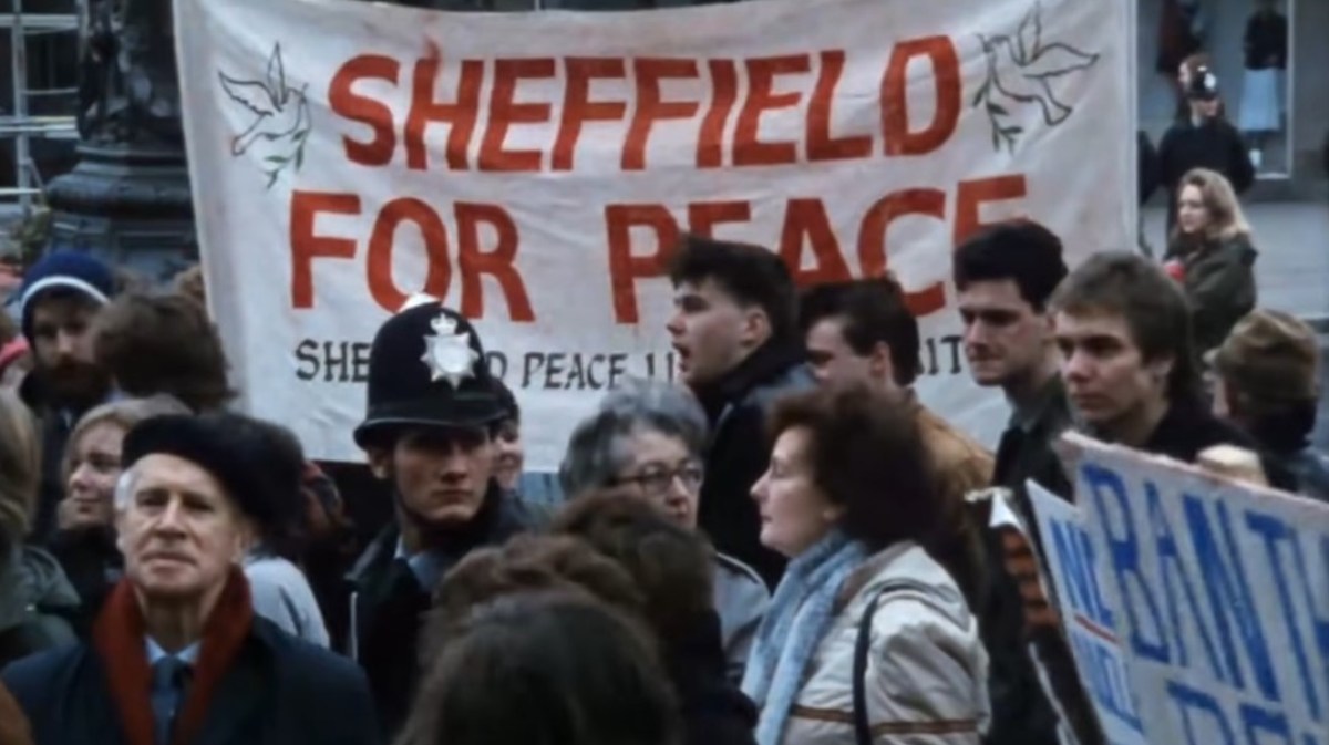 A British crowd of protestors hold a white sign reading, “Sheffield for Peace” in ‘Threads.’