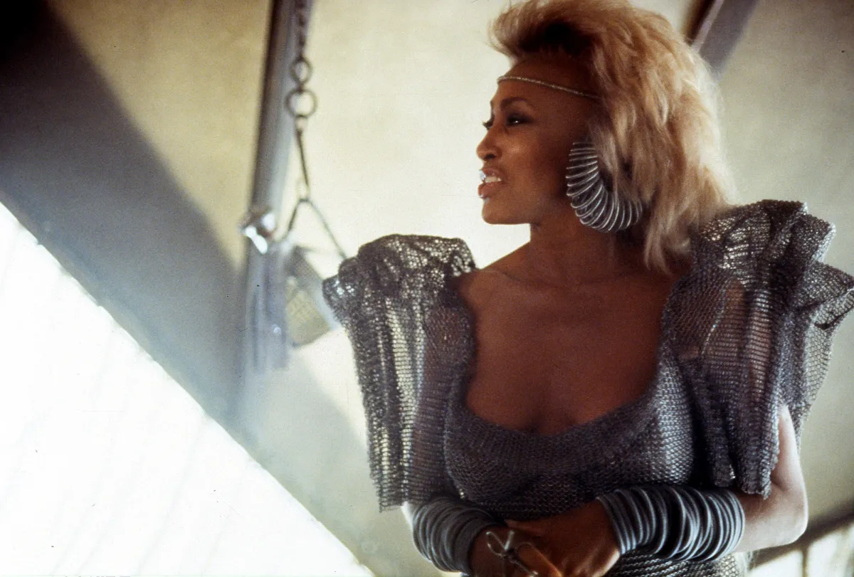 Tina Turner poses as Aunty Entity, wearing a mesh dress with puffed sleeves.