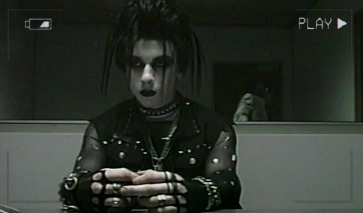 Image of a goth teenage boy sitting in an interrogation room in a scene from V/H/S/85. The image looks grainy as if off of a VHS tape, and has a low battery icon in one corner and says "PLAY" in the other. 
