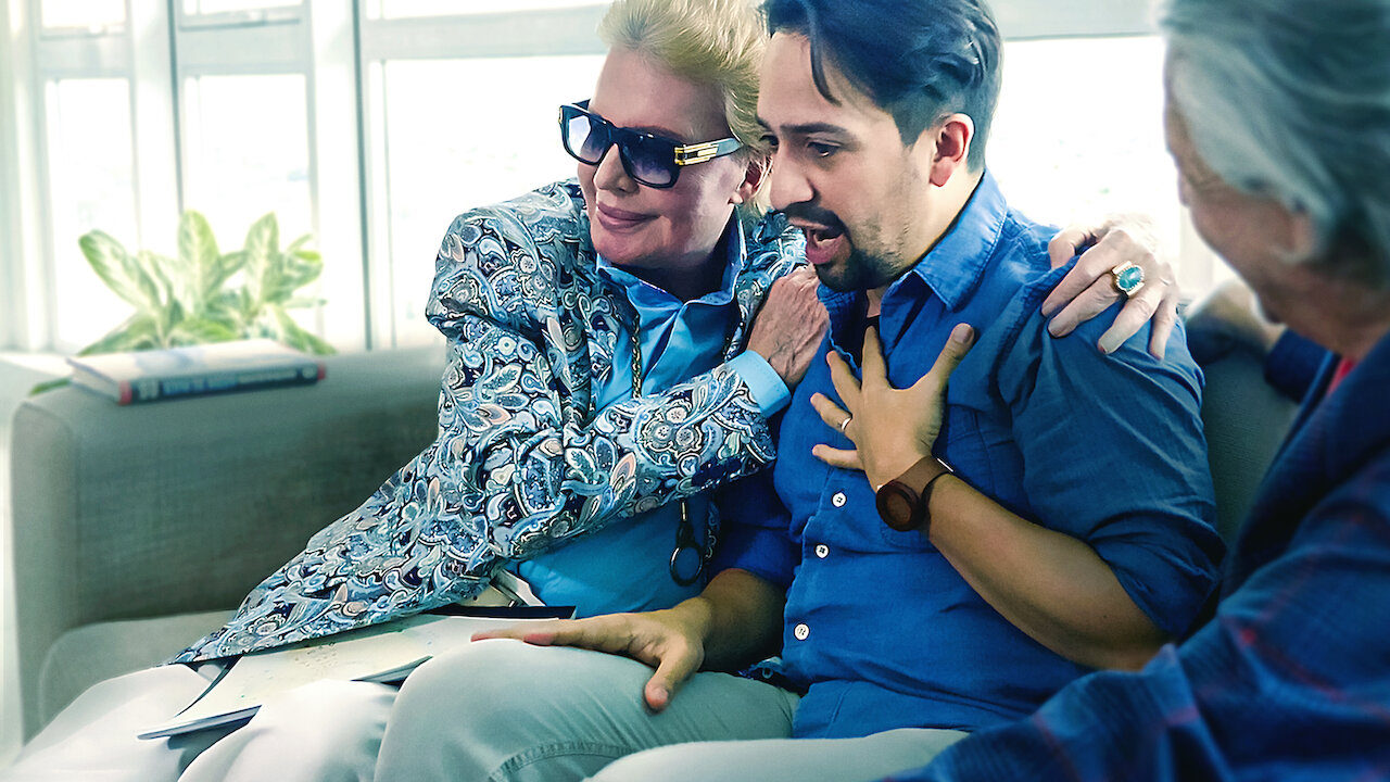 Walter Mercado sitting on a couch with his arms around Lin-Manuel Miranda in a scene from Netflix's 'Mucho Mucho Amor: The Legend of Walter Mercado.' 