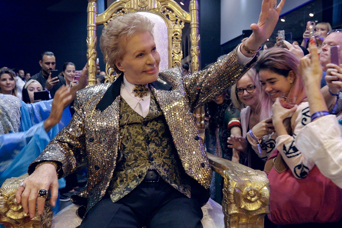 Image of Walter Mercado being brought into a room on a gold throne into a group of cheering fans in a scene from the Netflix documentary 'Mucho, Mucho Amor: The Legend of Walter Mercado.' Walter is a white Puerto Rican man with short wavy blond hair and is wearing a glittery gold blazer over a white button down with a large gold brooch at his throat, a black vest with a gold leaf pattern, and black pants. He's waving to the crowd.
