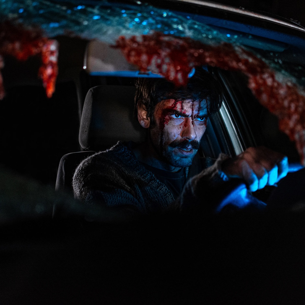 Ezequiel Rodriguez in a scene from 'When Evil Lurks.' He is a white Latino with shaggy dark hair and a black mustache. He's sitting in the driver's seat in a car with a crashed out, blood-splattered windshield. HIs face is covered in blood. 