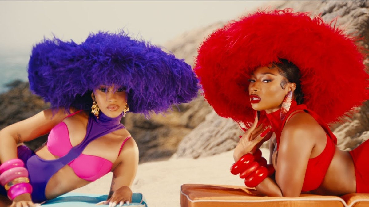 Cardi B and Megan Thee Stallion on a beach in extravagant colorful sun hats in a still from the "Bongos" music video