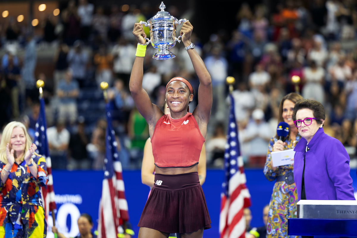 Young Black tennis champ Coco Gauff holds a trophy above her head and grins.