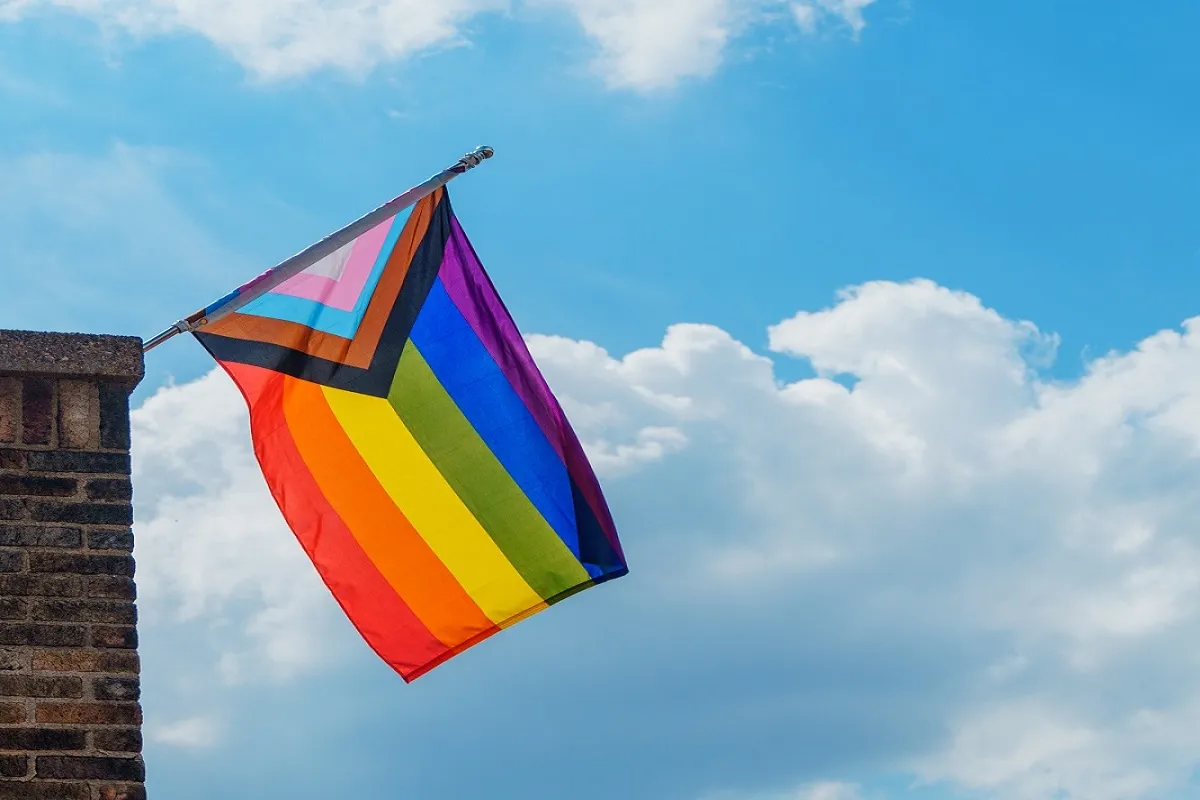 A photo of single Progress Pride Flag, mounted on a chimney to the left, with a blue sky and clouds in the background.