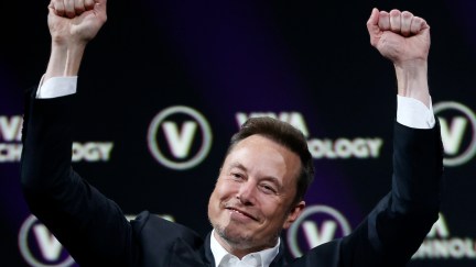 Elon Musk holds his fists above his head in mock triumph.
