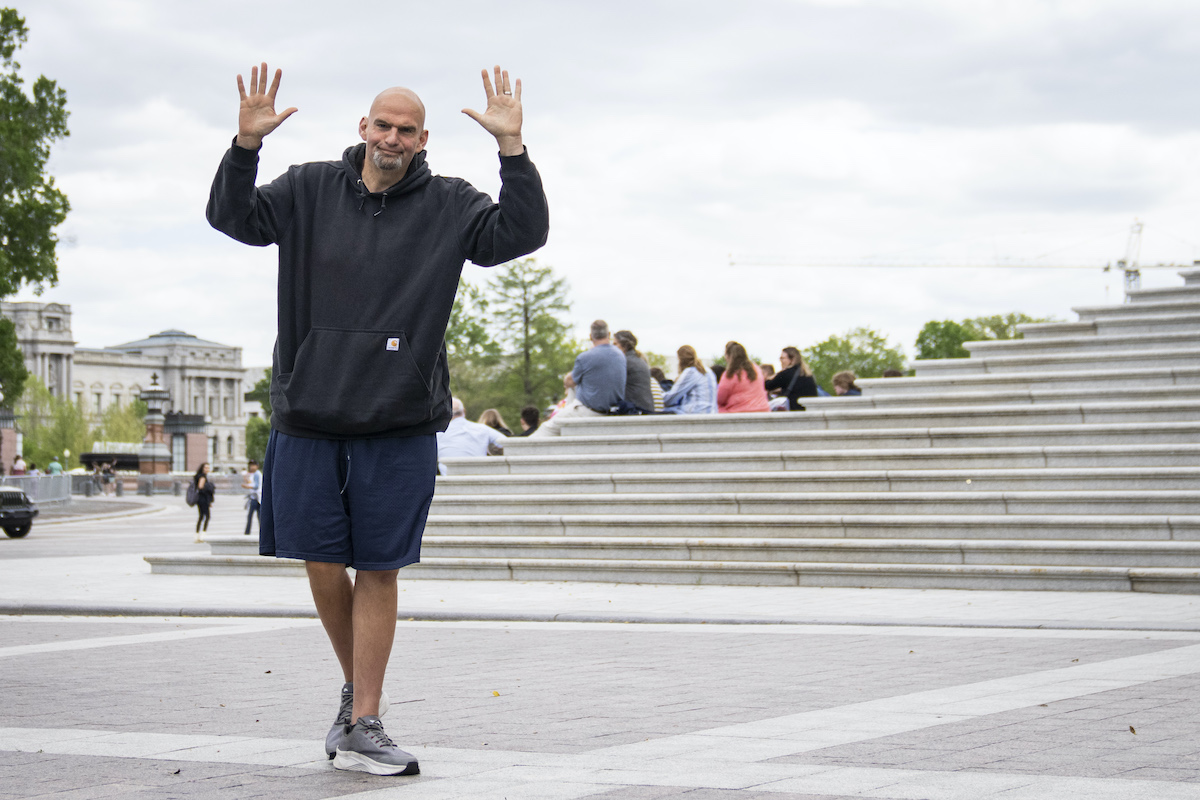 John Fetterman wears a hoodie and basketball shorts, and waves with both hands.