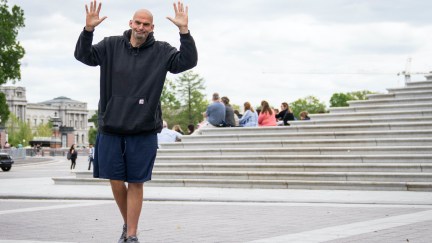 John Fetterman wears a hoodie and basketball shorts, and waves with both hands.
