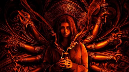 A girl stands in dim red lighting with Hindu deity-like arms spreading behind her. She holds a jar and stares at the camera.