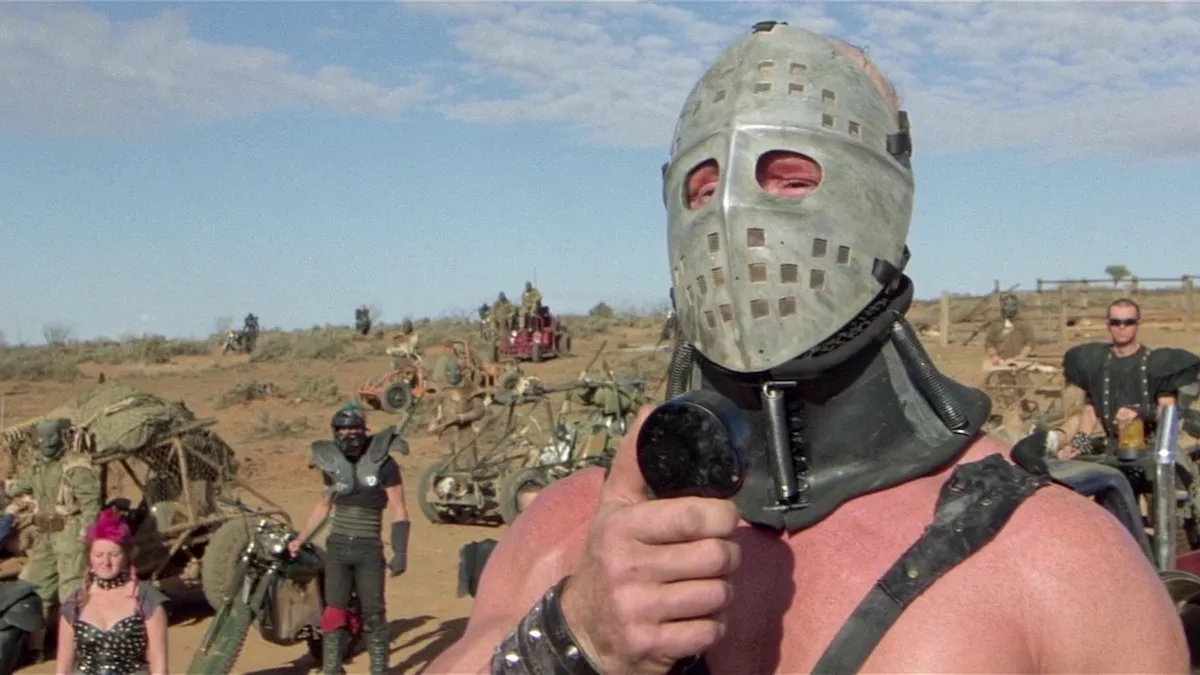 A shirtless man in a hockey masks speaks on a hillside, with warriors behind him.