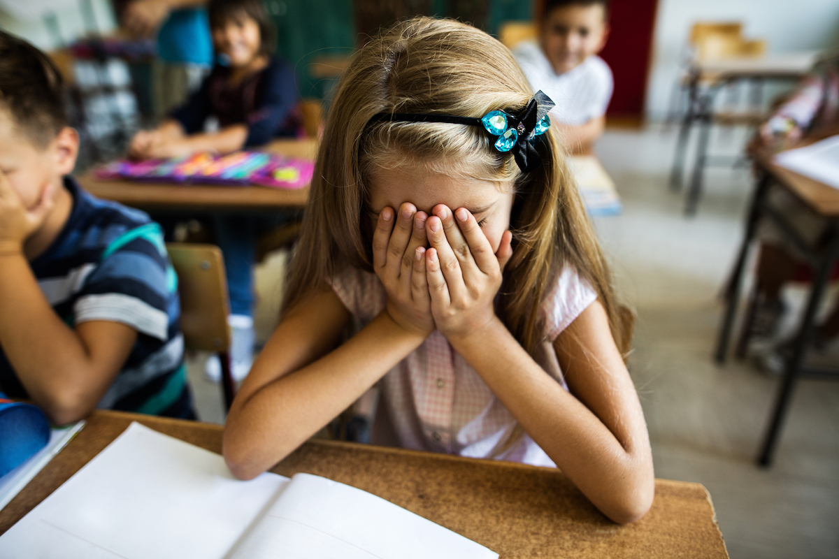 Little girl in the classroom feeling stressed and covering her face with hands while sitting at the desk.